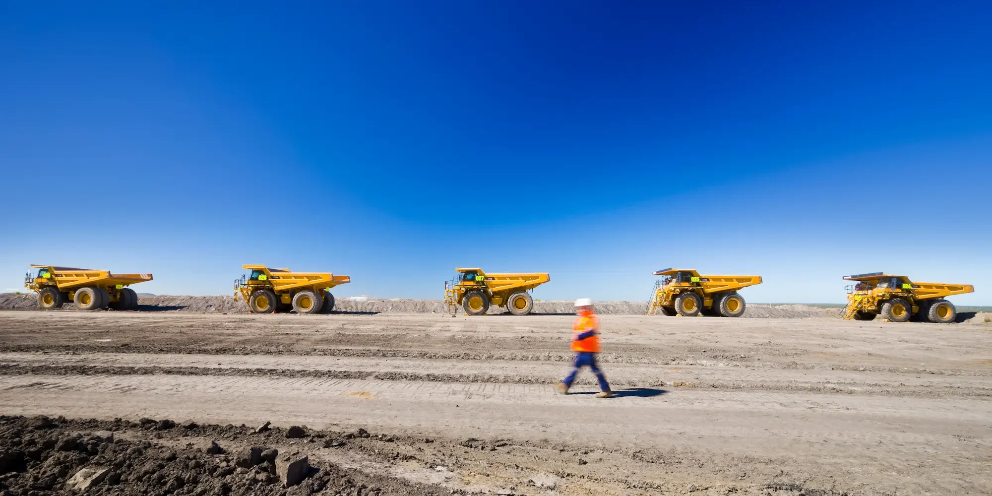 Worker walking alongside line of dump trucks at job site.