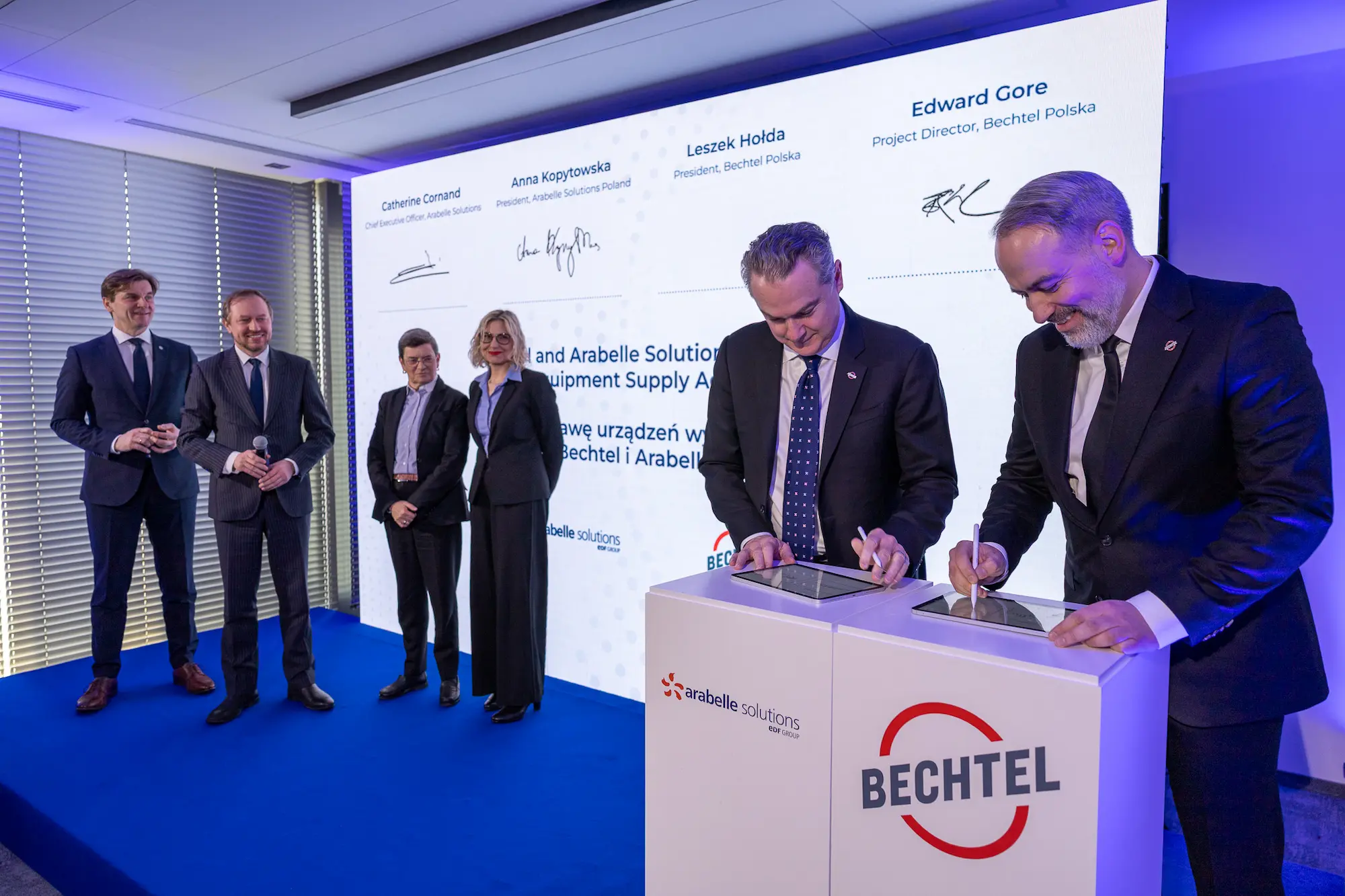 Image of two men signing ceremonial documents as the rest of their team looks on from the side. Backdrop is a large LED screen.